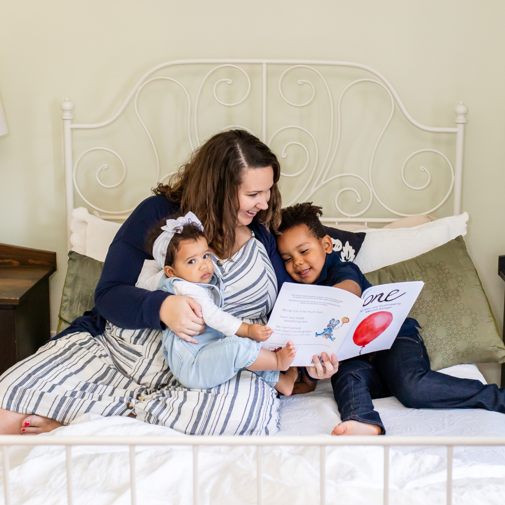 Mother and two children snuggling in bed reading story book, "One: A Birthday Book".